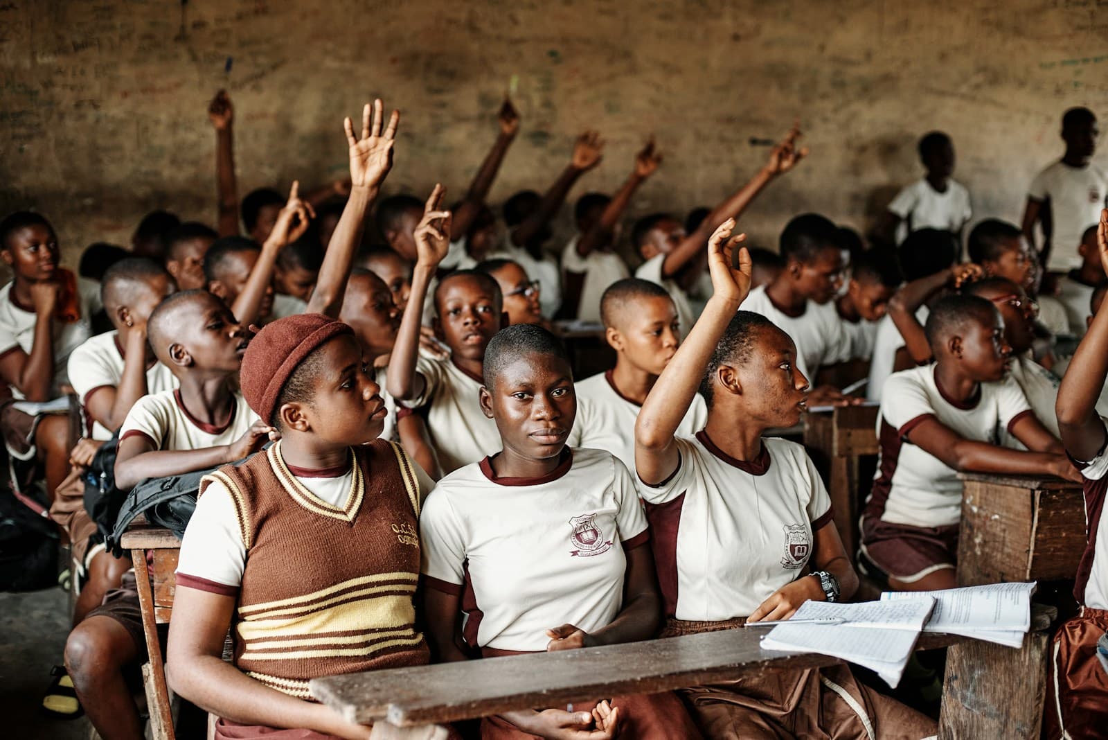 A classroom of students in school uniform, several with their hands raised to answer.