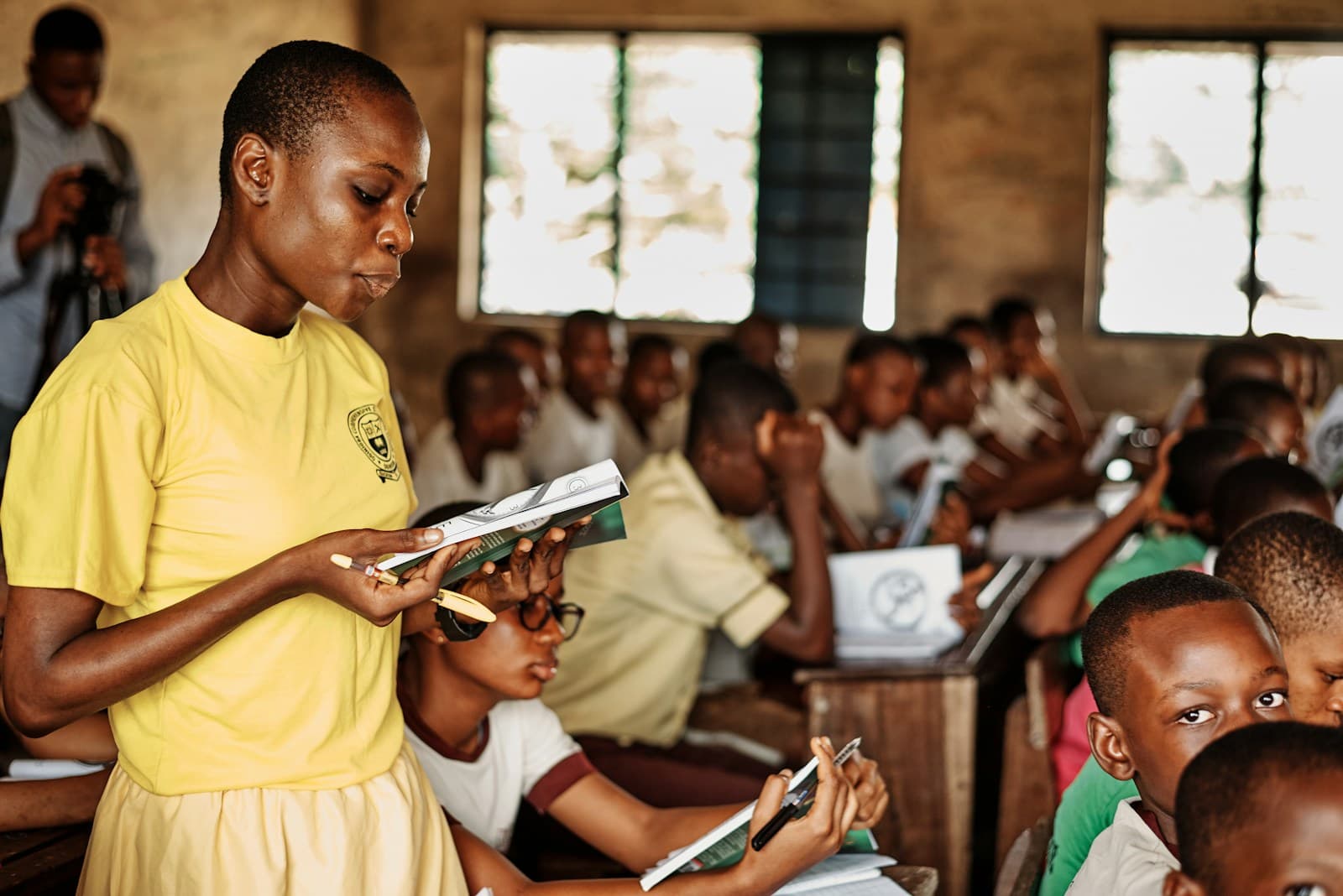 A secondary school student in a yellow uniform reads aloud from a textbook while classmates follow along at their desks.